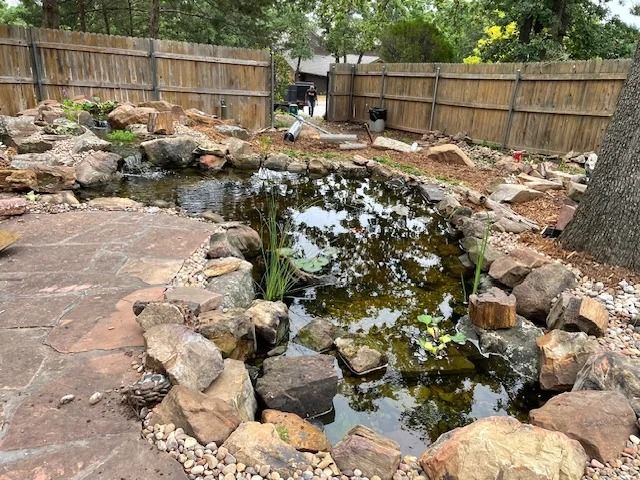 Backyard pond with rocks, shallow water, and a wooden fence surrounded by trees