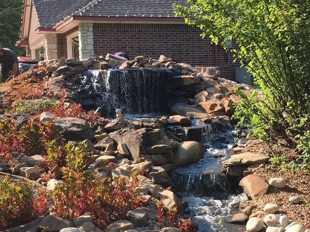 Rock waterfall garden beside a brick house with red and green shrubs