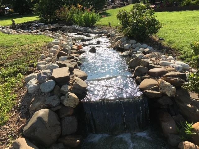 Stone-lined stream waterfall in a sunny park with green grass and shrubs