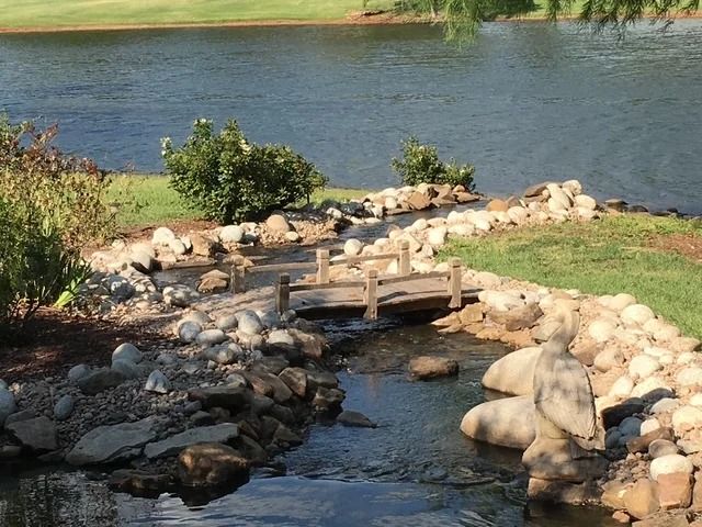 Rock-lined garden stream with a small wooden bridge flowing into a lake or pond