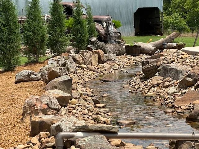 Rock-lined backyard stream with flowing water and small evergreen trees along a fence