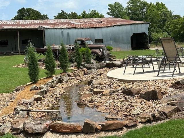 Backyard pond and stone landscaping beside a patio with chairs near a rustic outbuilding