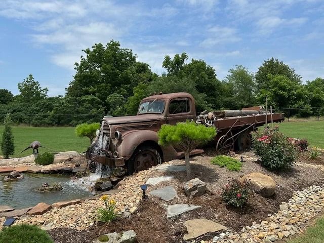 Old rusty truck partly submerged in a landscaped pond with rocks and greenery.
