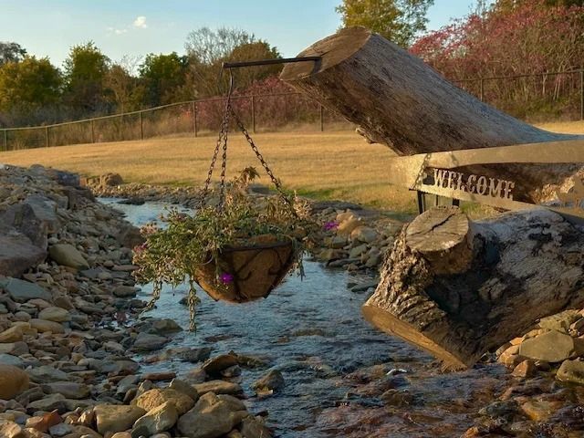 Rustic metal crane hoists a hanging basket over a shallow rocky stream in a rural field