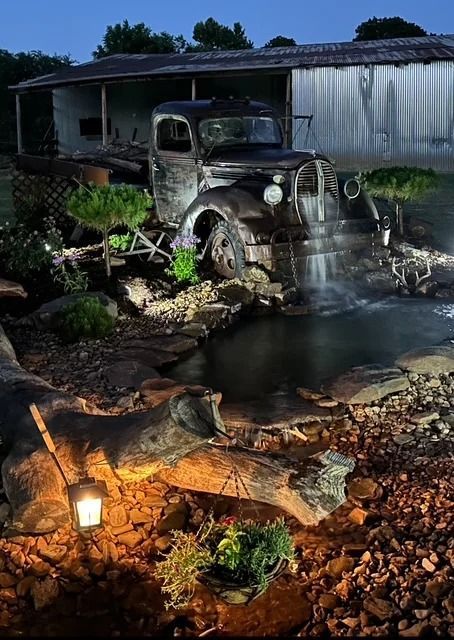 Old rusty truck beside a small waterfall at night in a landscaped garden with rocks and lantern light