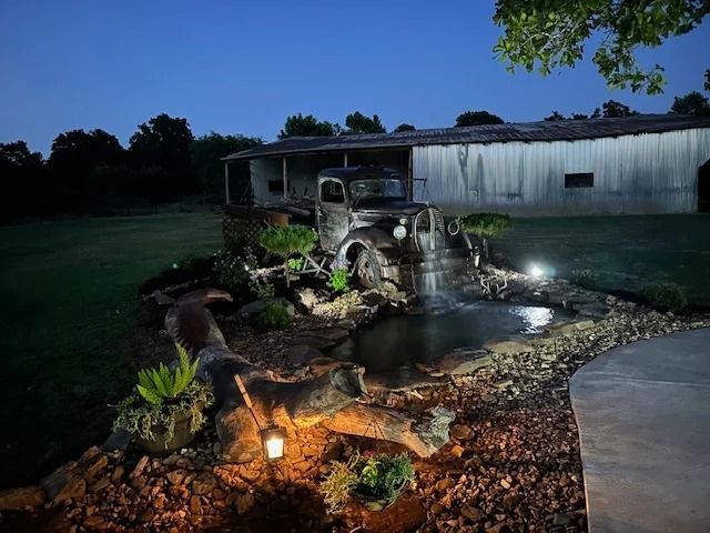 Nighttime garden pond with small waterfall, rocks, and lights beside a house