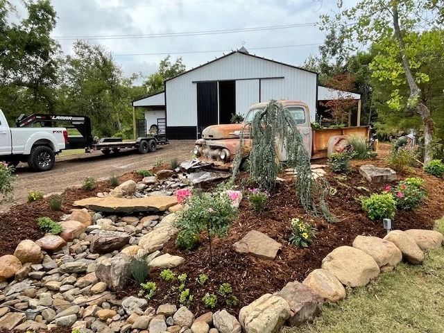Gravel garden with rocks and flowers beside a white barn and parked pickup trucks