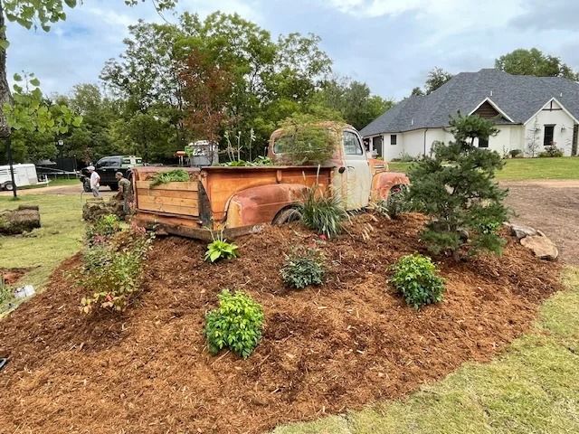 Rust-colored vintage truck bed in a mulched garden bed with shrubs and a house in the background