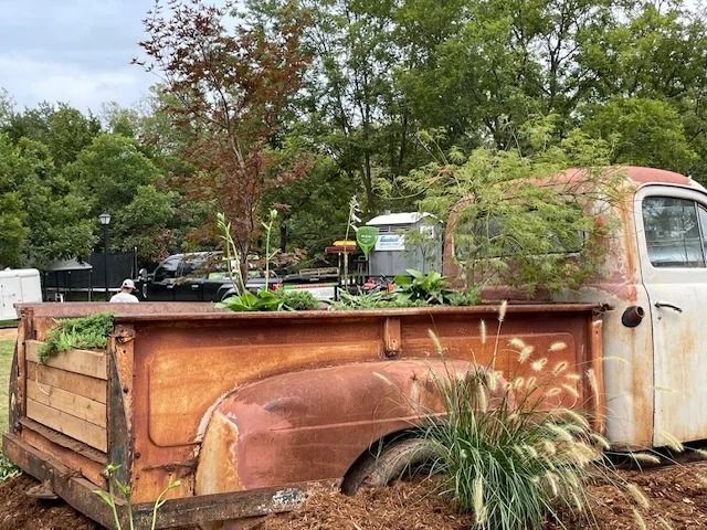 Rusty pickup truck planter with greenery in a garden setting, surrounded by trees and shrubs.