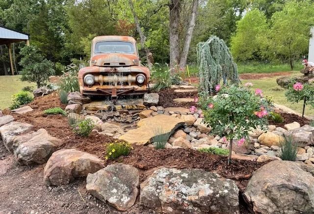 Rusty vintage truck in a landscaped garden with rocks, flowers, and a small pond on a rainy day
