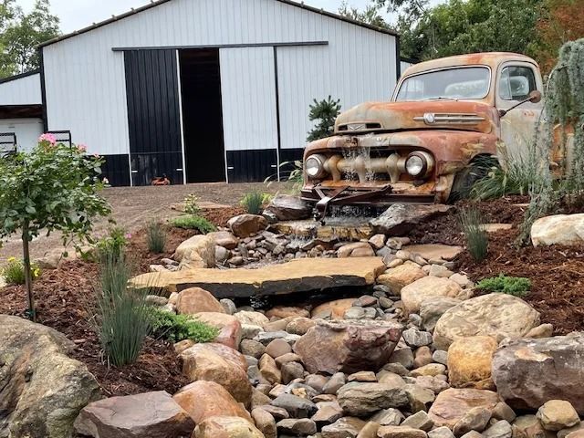 Rusty vintage truck beside a white barn with a stone-lined dry creek bed in the foreground