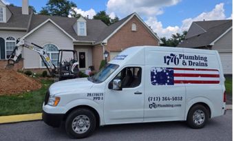 A plumbing and drains van is parked in front of a house with an excavator in the yard