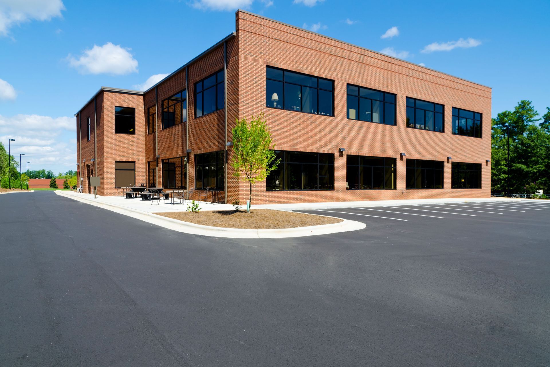 A modern two-story brick office building with dark-tinted windows, situated next to an asphalt parking lot under a blue sky.