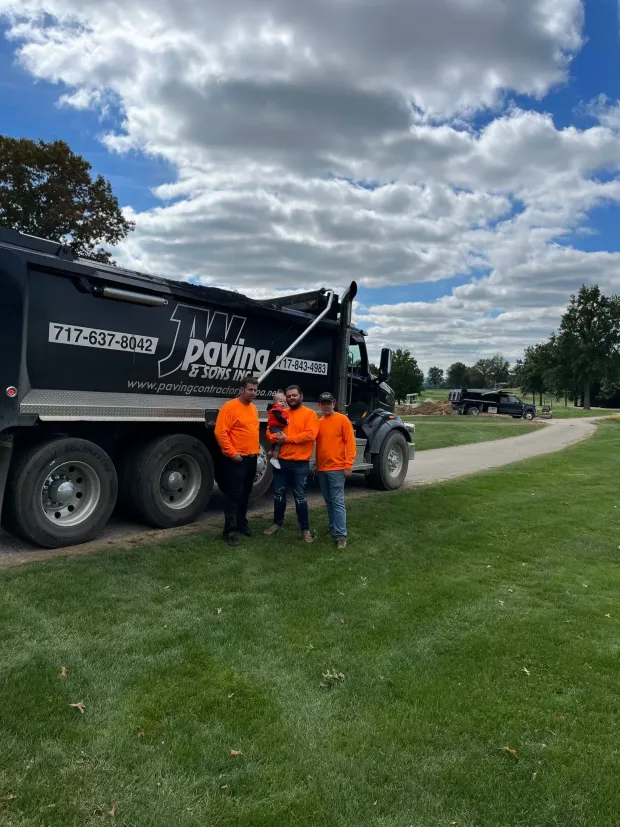 Three people in orange high-visibility shirts stand next to a black dump truck labeled “Bauer Paving” on a grassy path.