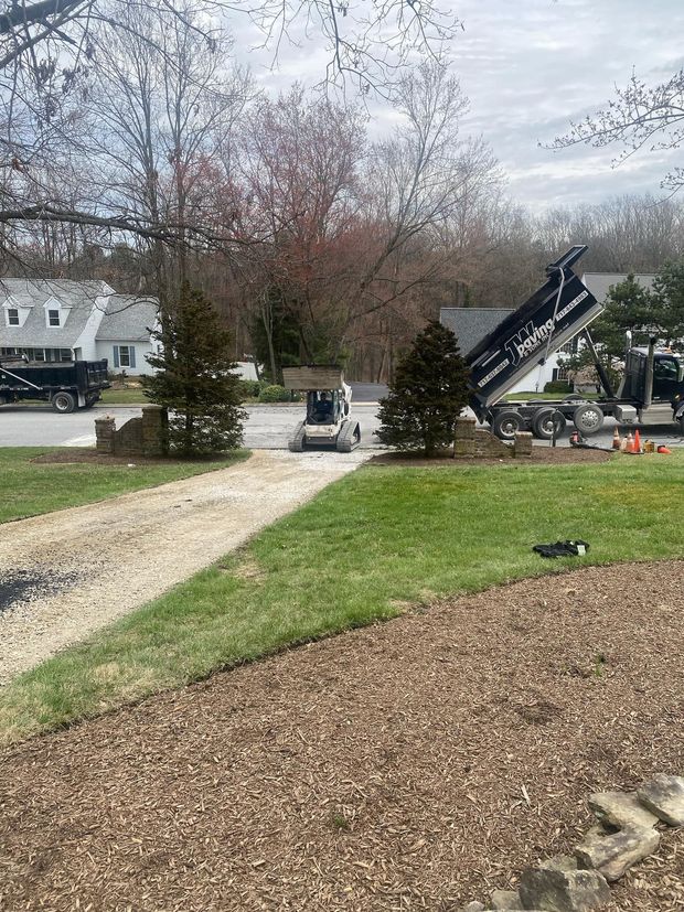 A construction site with a skid steer, a dump truck, and gravel piles in a suburban residential driveway.