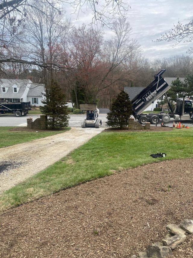 A construction site with a skid steer, a dump truck, and gravel piles in a suburban residential driveway.