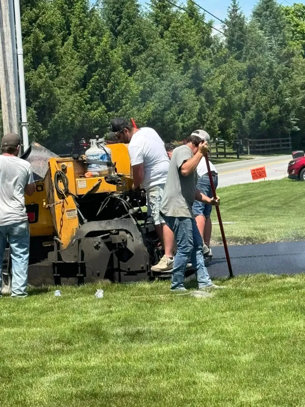A crew of four people uses a yellow paving machine to lay fresh asphalt on a sunny day in a grassy area.