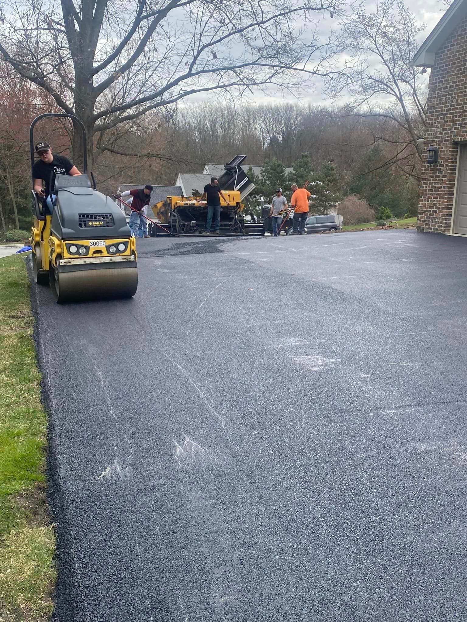 Construction crew paving a driveway with a yellow asphalt roller and machinery in front of a brick house.