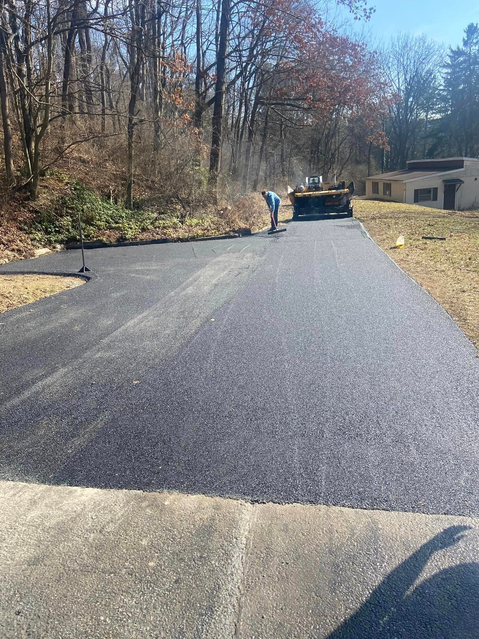 A person in blue jeans stands on a freshly laid black asphalt driveway leading to a building near a wooded area.