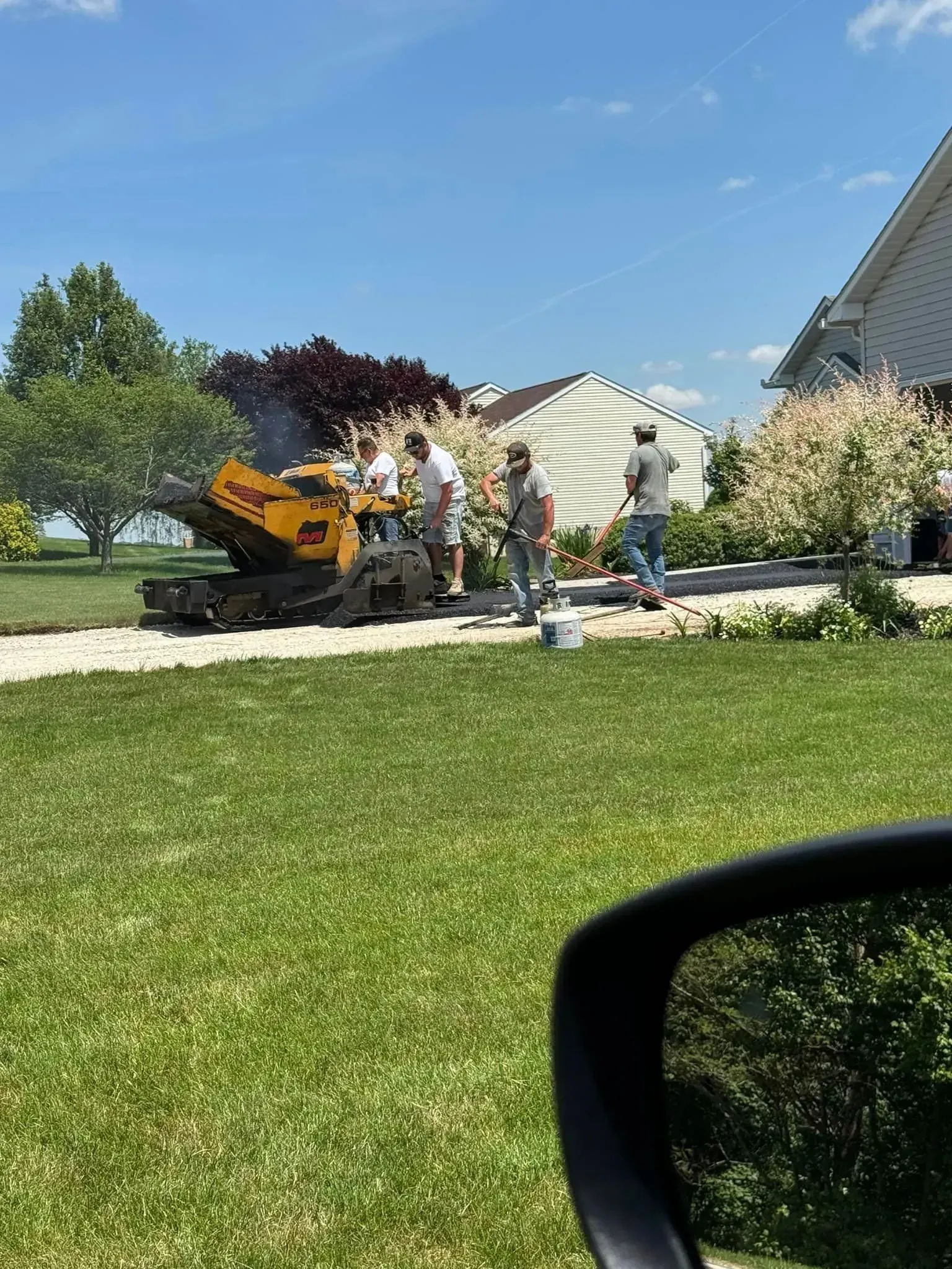 Workers use a yellow construction machine on a sunny day in a residential yard near houses and trees.