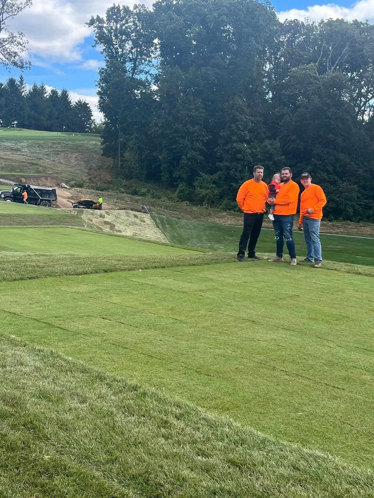 Three people in matching orange shirts stand on a freshly landscaped lawn with wooded hills in the background.
