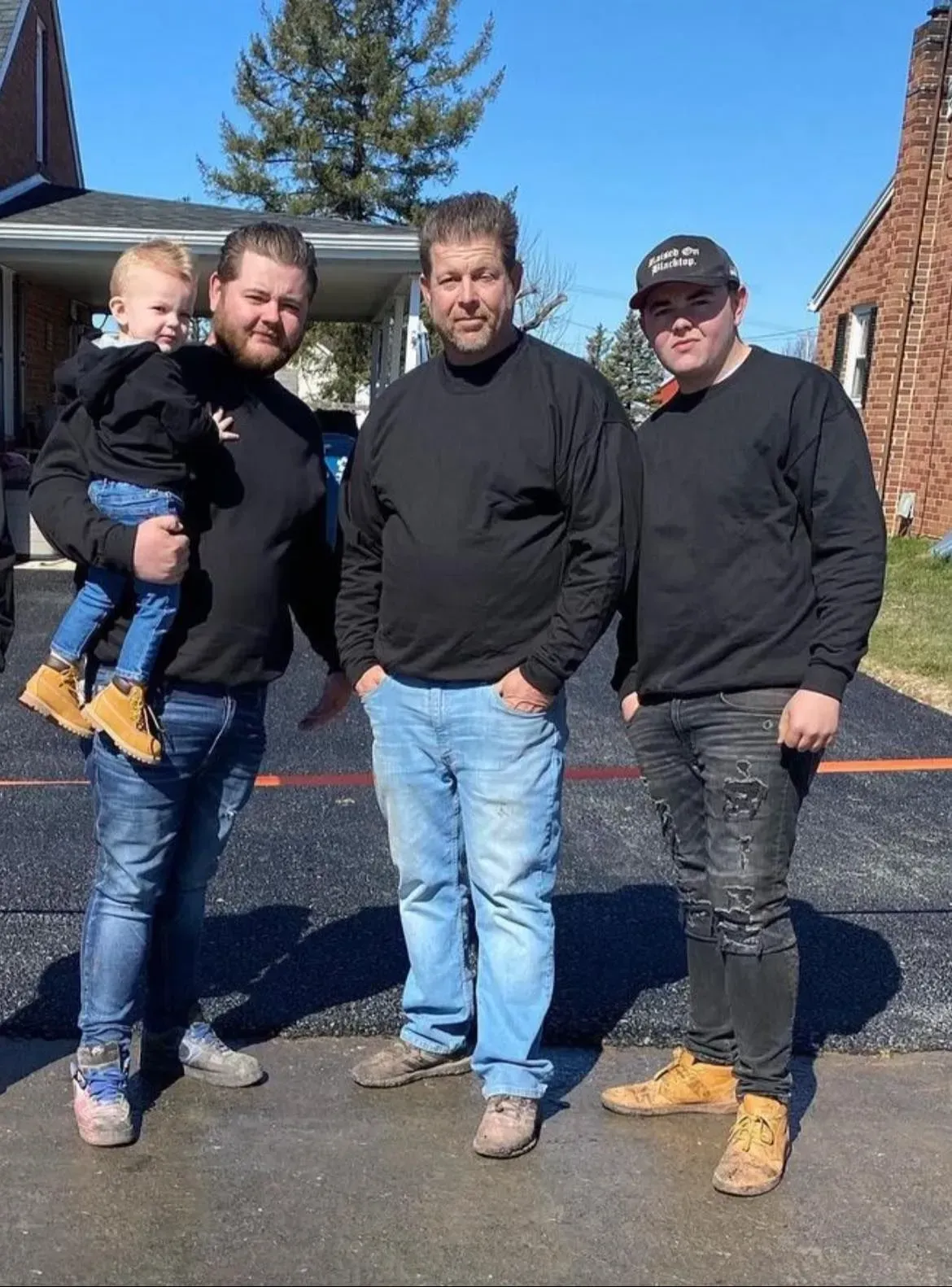 Three men and a child stand together on a paved driveway in front of a house on a sunny day.