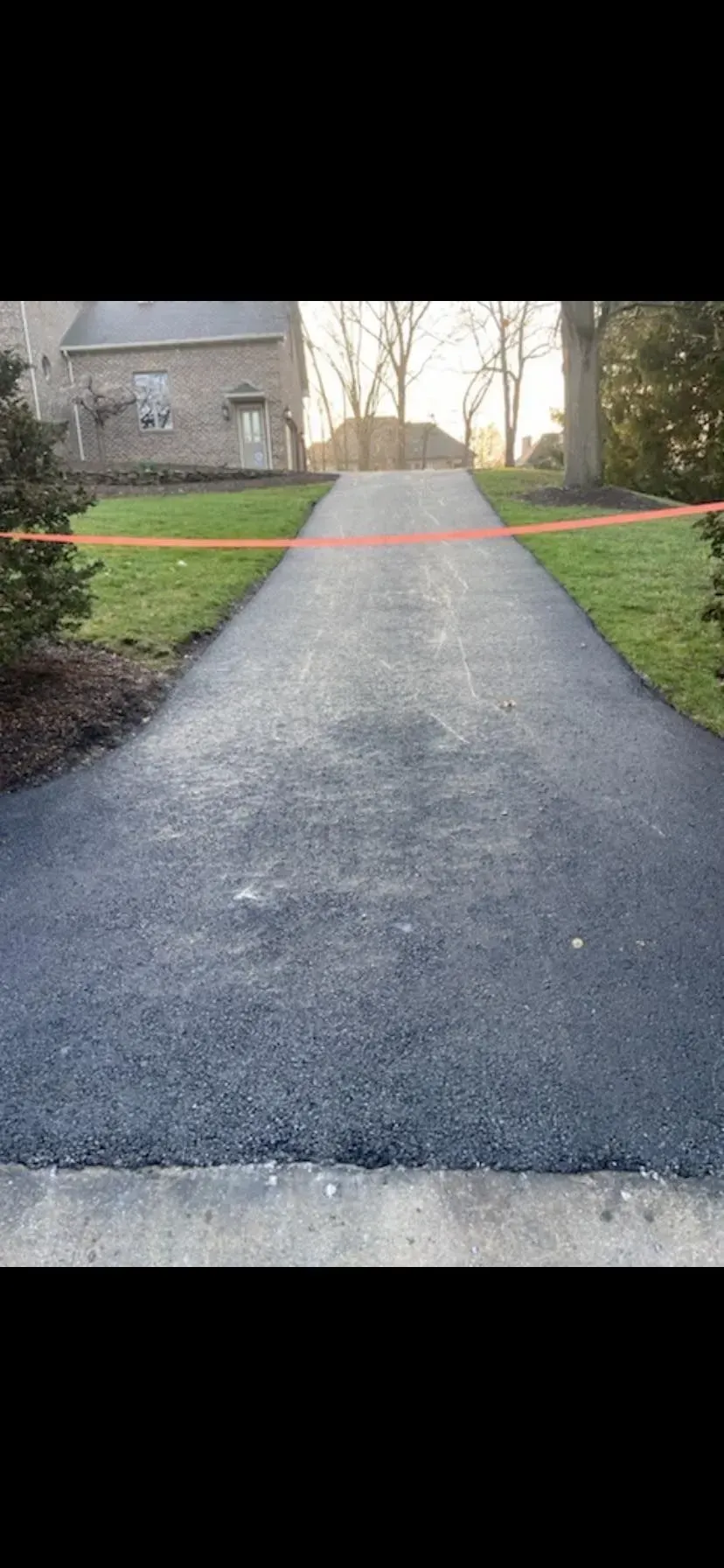 A newly paved asphalt driveway leads toward a brick house, blocked by an orange caution tape.