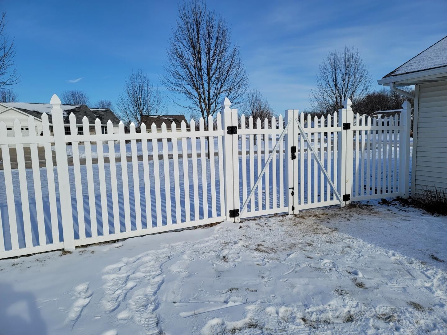 A white picket fence with a gate in the snow next to a house.