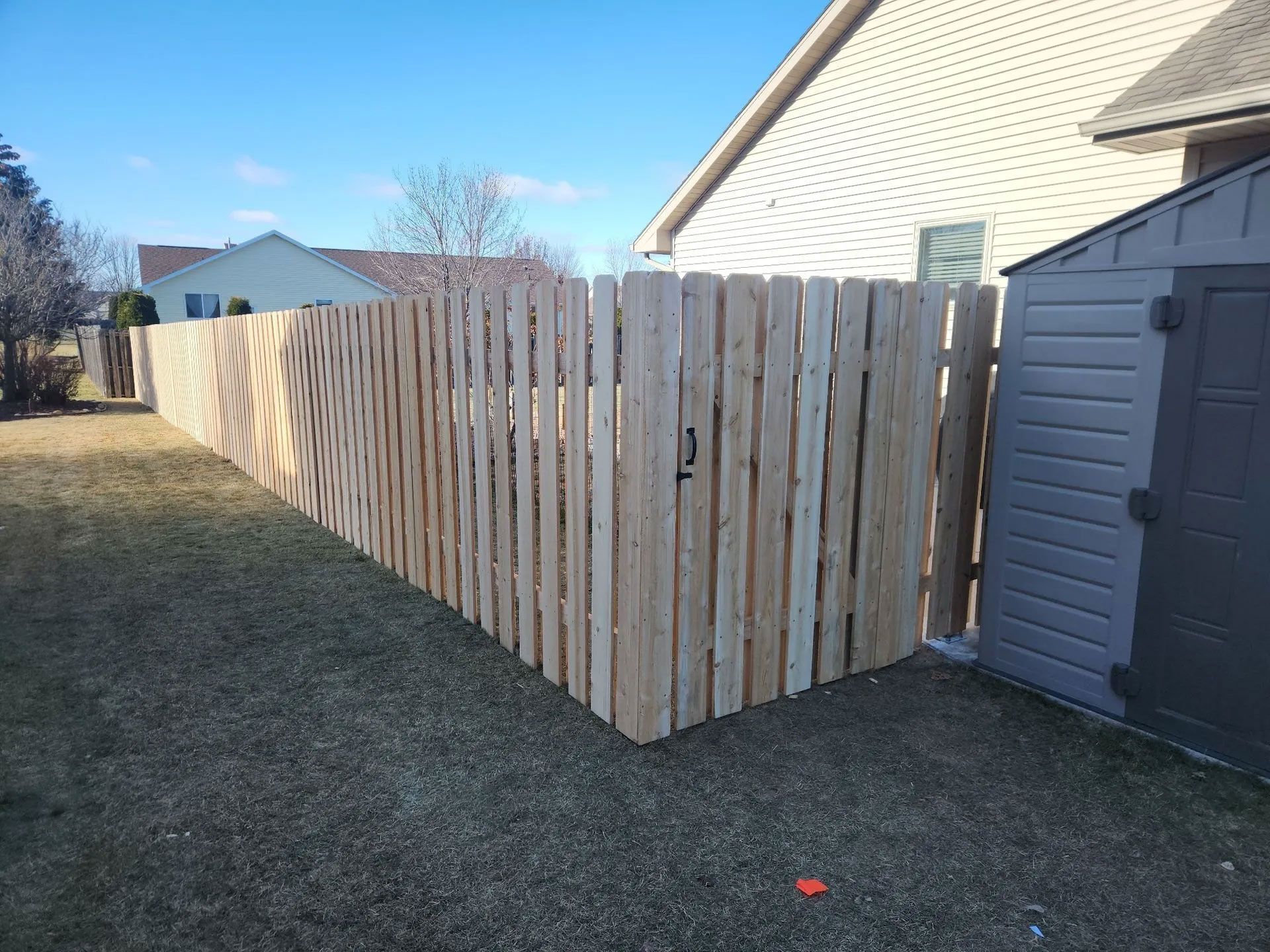 A wooden fence encloses a yard with a shed on the right. Clear skies and adjacent houses are visible.