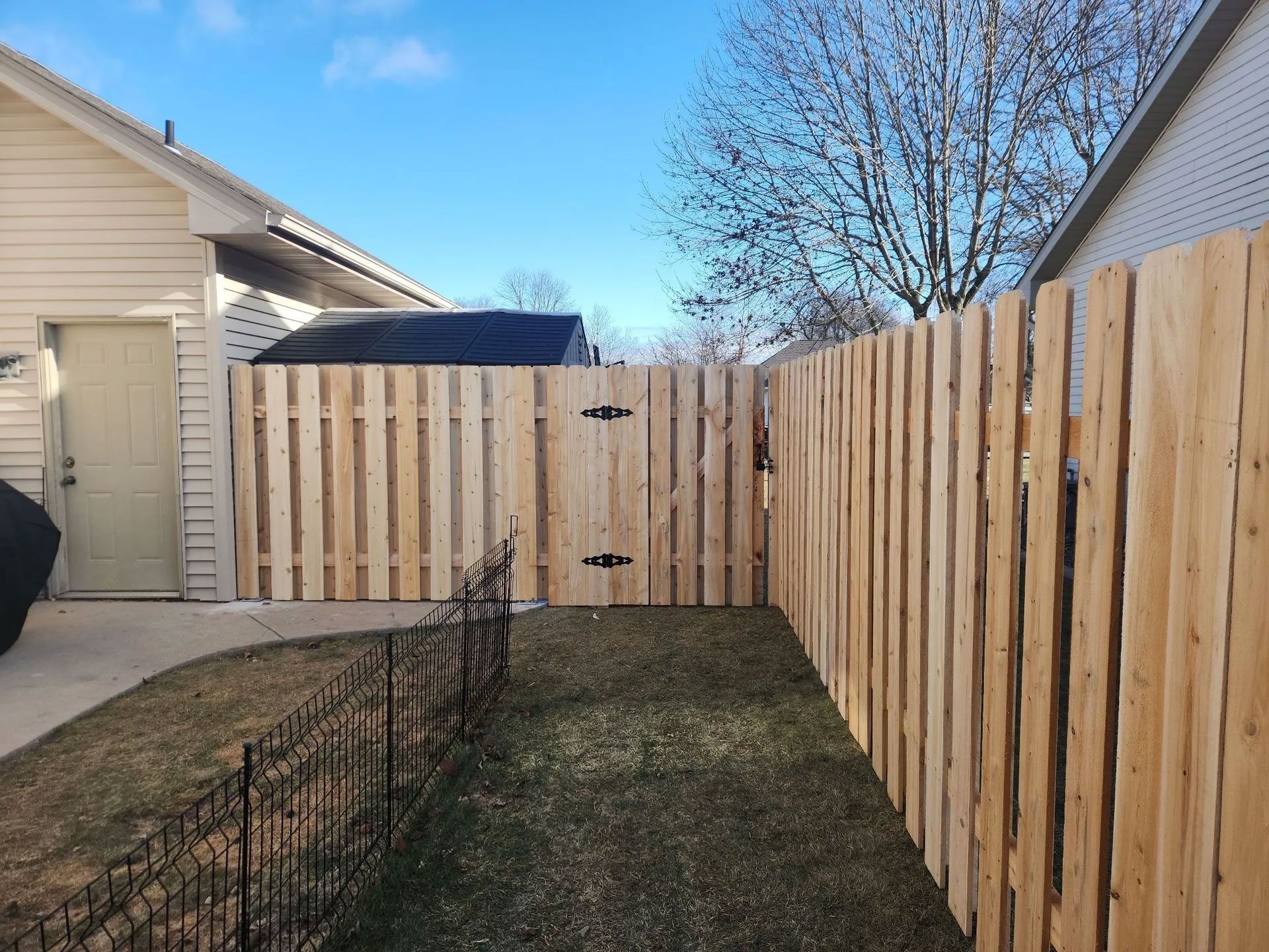 A wooden fence with a gate encloses a yard next to a garage and a house on a sunny day.