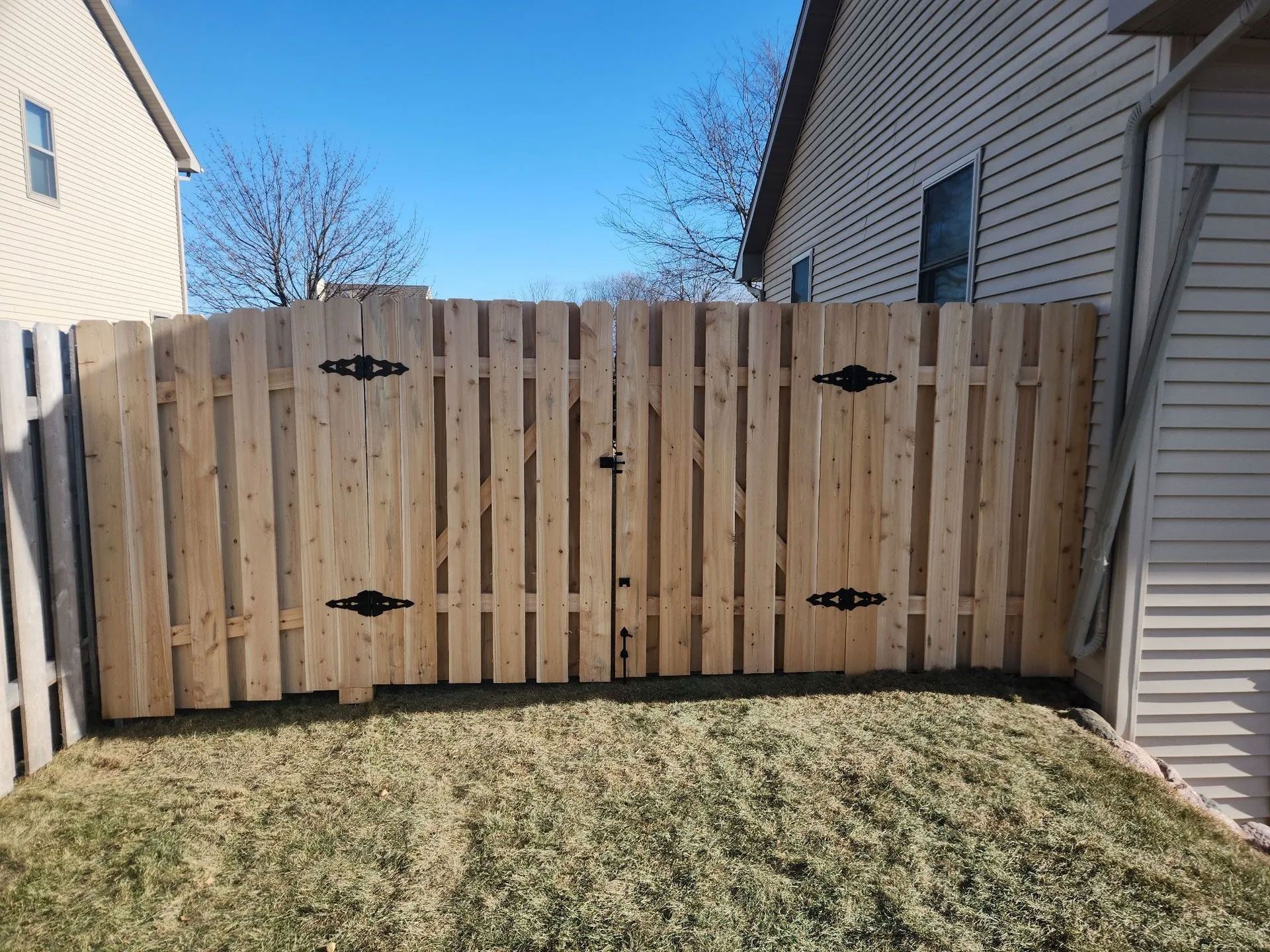 Wooden fence with a gate in a backyard setting. The fence and gate have black decorative hardware.