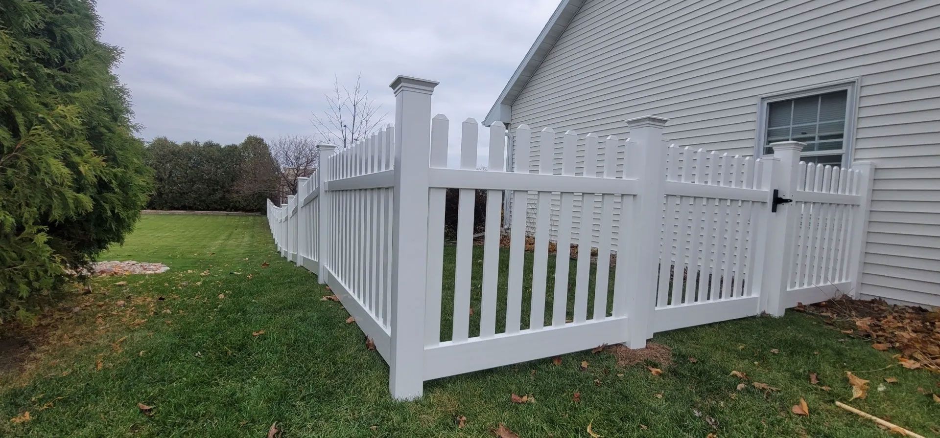 A white picket fence curves around a grassy yard, next to a house with white siding and a window.