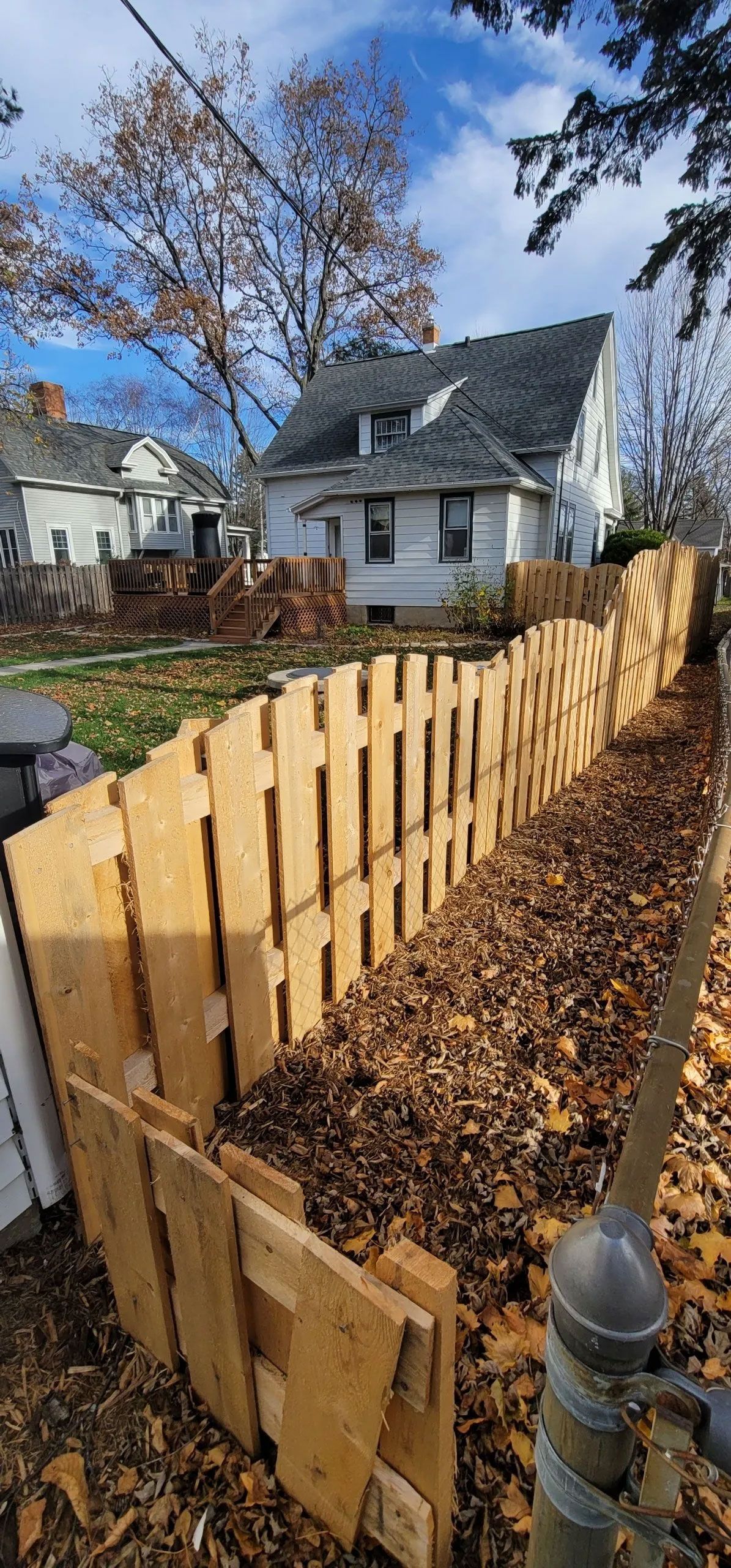 A wooden fence filled with leaves in front of a house. The sky is blue with clouds.