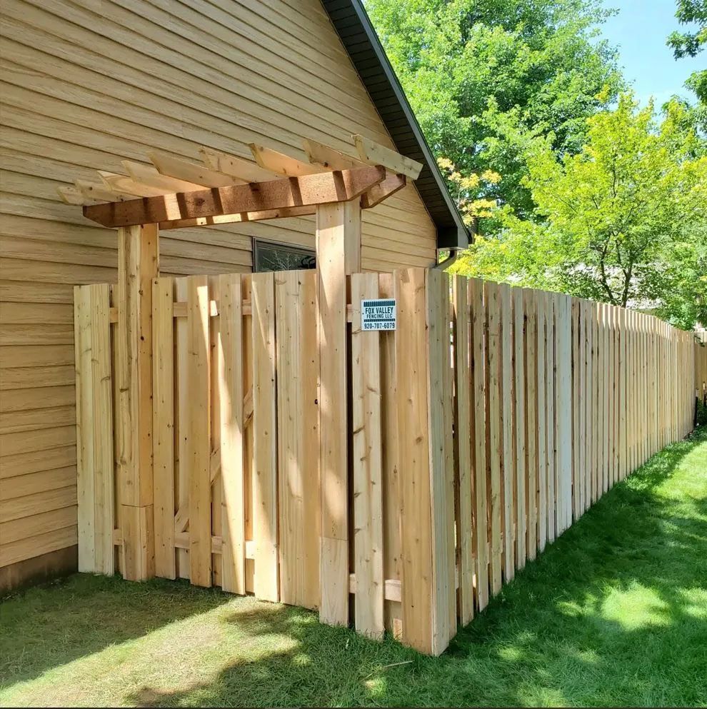 A wooden fence surrounds a house with a small pergola over a gate, all made of light-colored wood. Green grass and trees are in the background.