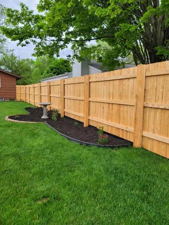 A wooden fence lines a green lawn with a flower bed and bird bath along its base. A tree and a building are in the background.