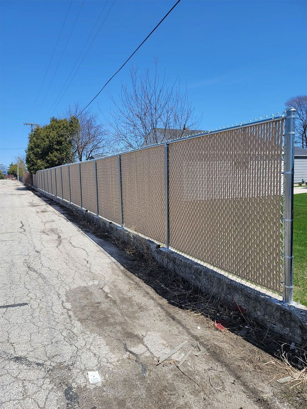 Chain-link fence with tan privacy slats alongside a cracked, gray alley on a sunny day.