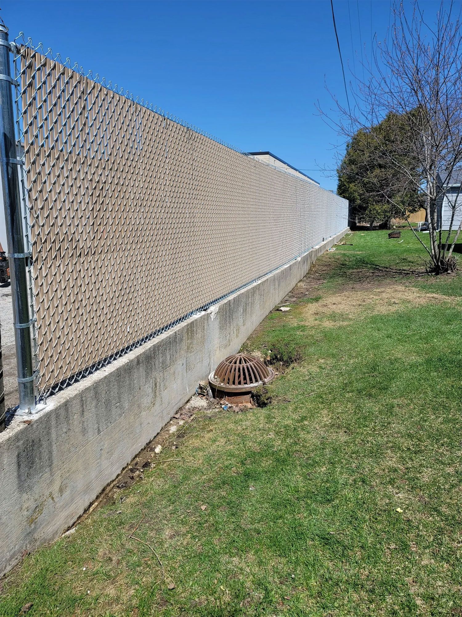 Chain link fence with tan privacy slats atop a concrete retaining wall, next to a grassy lawn and a storm drain.