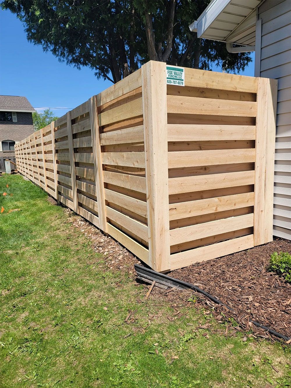 A wooden fence constructed of horizontal planks encloses a yard, with the fence appearing light brown and set in a residential area with grass.