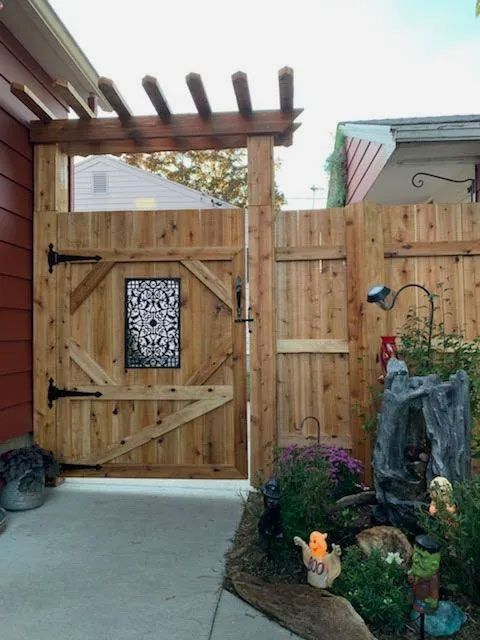 Wooden gate and fence with a pergola, leading into a garden with flowers, a small tree stump, and decorative lights.