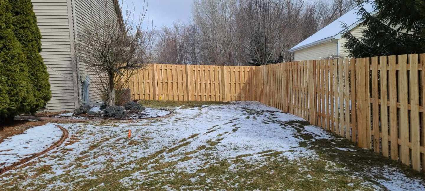 A wooden fence surrounds a backyard covered in snow. A house with a snowy roof is visible in the distance.