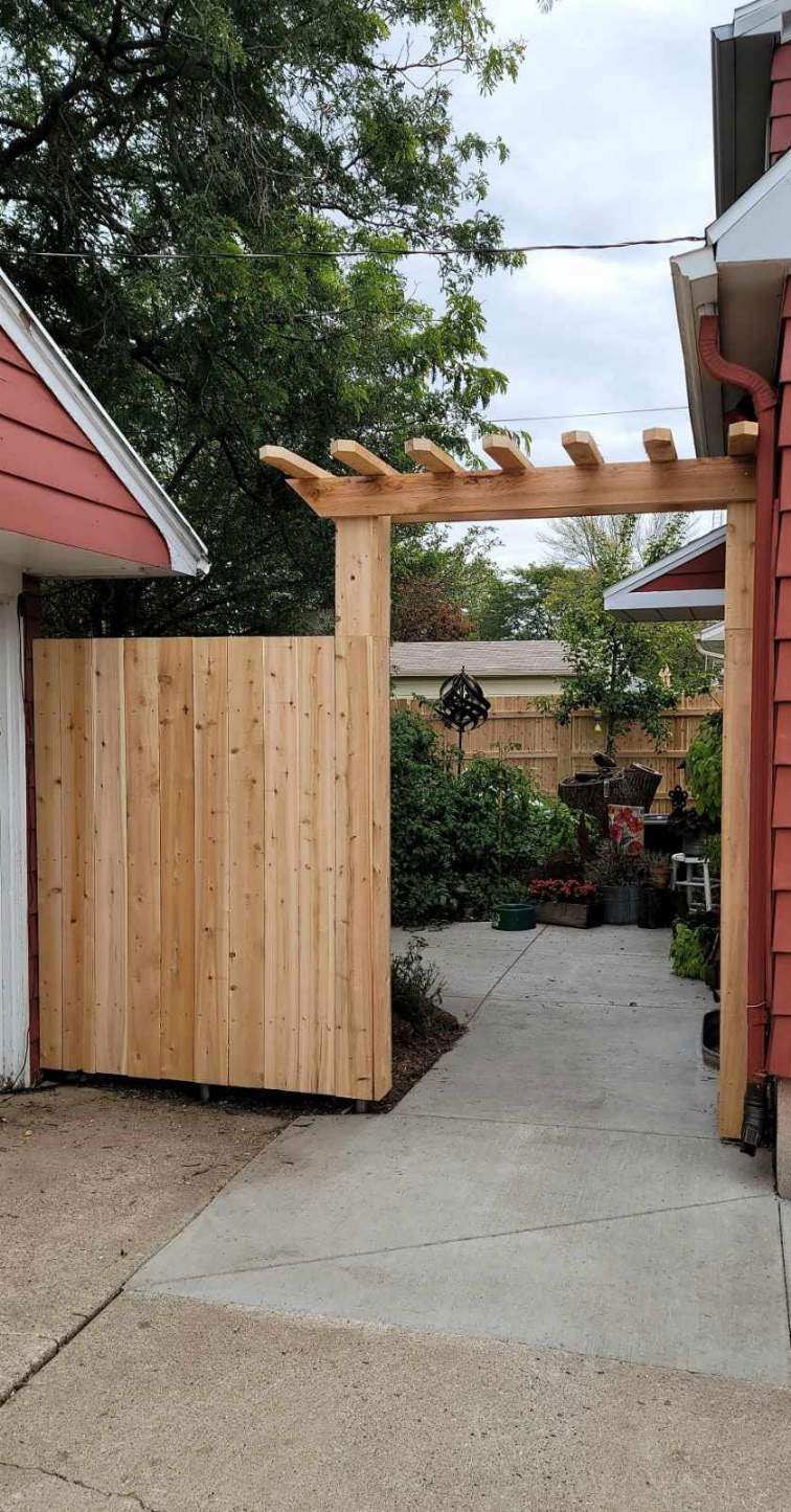 A wooden gate with an overhead trellis leads to a backyard. The gate and surrounding structure are light brown, and the setting is outdoors.