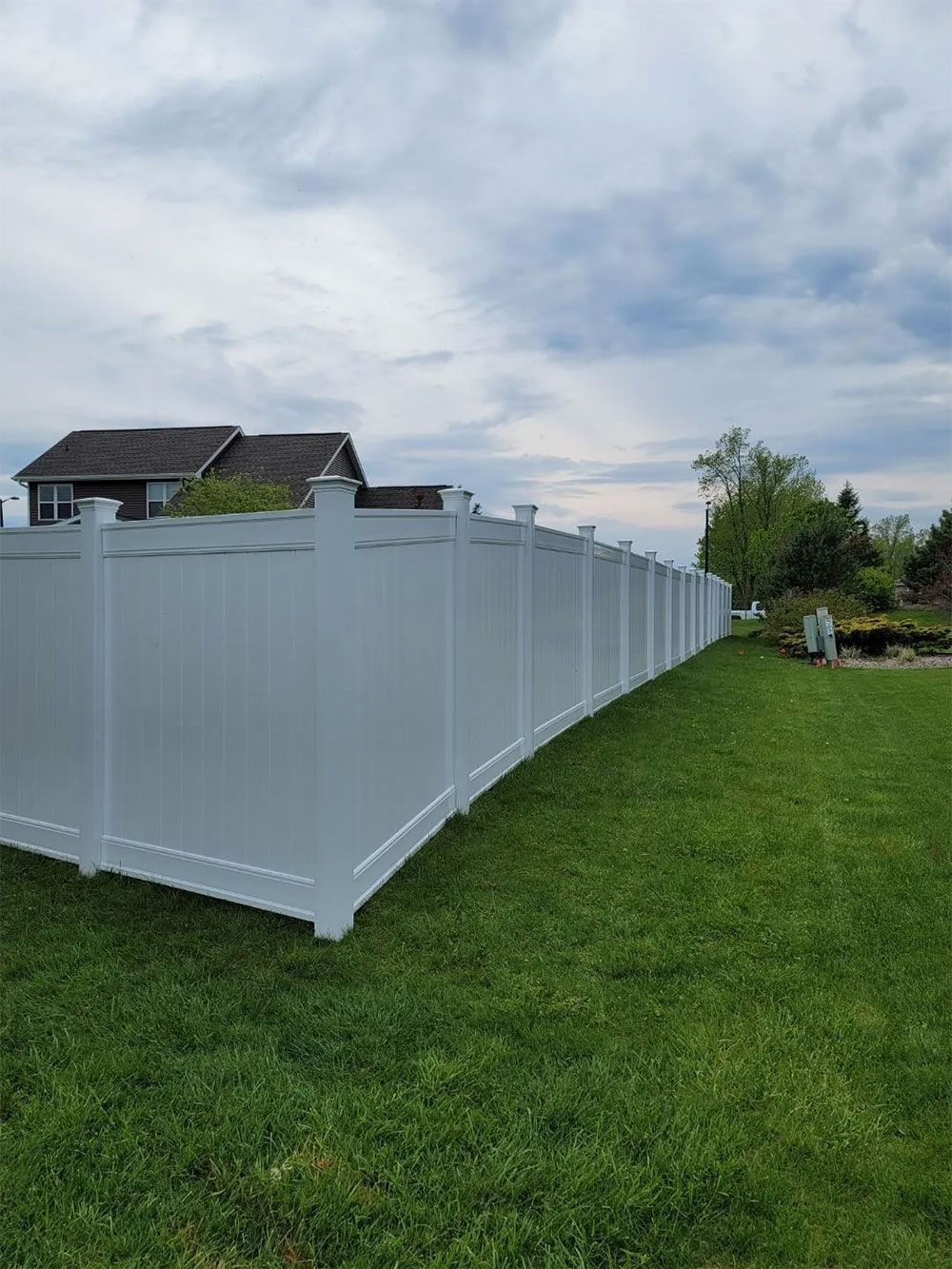 White vinyl fence in a grassy yard, curving along the property line on a cloudy day. A house is visible in the distance.
