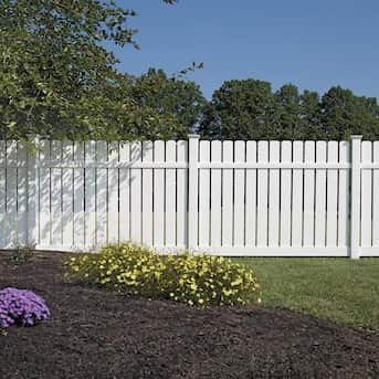 White picket fence bordering a yard with green grass and flowering bushes. Blue sky and trees are in the background.