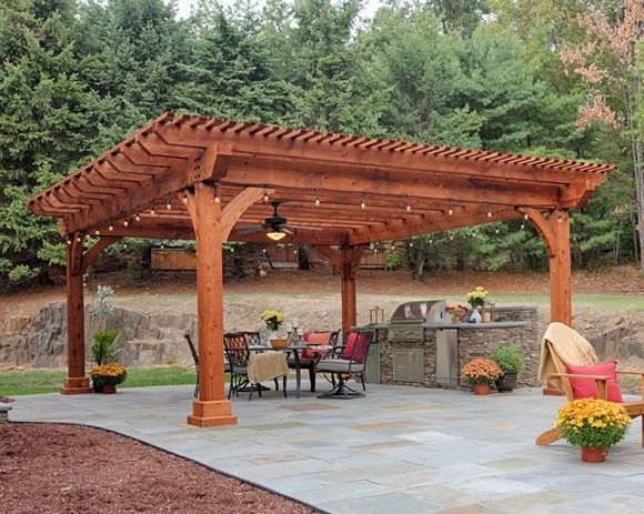 Wooden pergola over patio with dining set, grill, and seating; fall foliage backdrop.