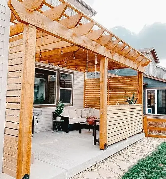 Wooden pergola with a seating area on a concrete patio. The structure has a slatted privacy wall.