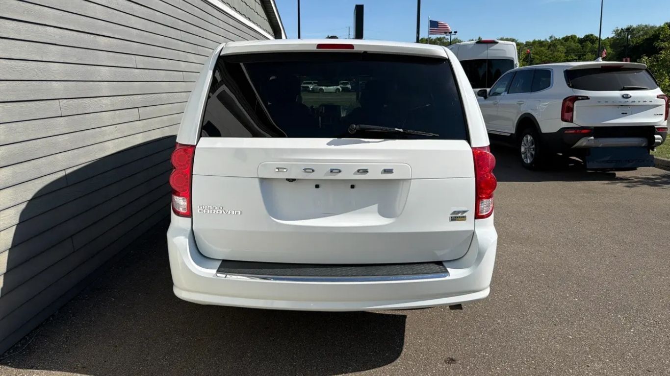 White minivan parked by a building wall in a lot, rear view with another SUV in the background