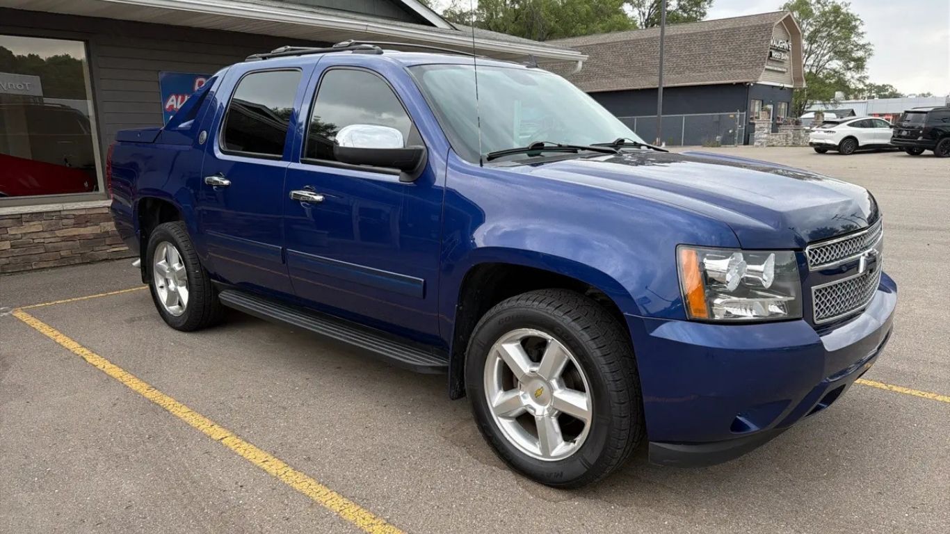 Blue pickup truck parked in a lot, angled front view, with yellow parking lines and buildings in the background