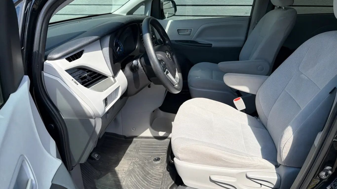Car interior with gray fabric seats and black steering wheel, viewed from the open driver’s door