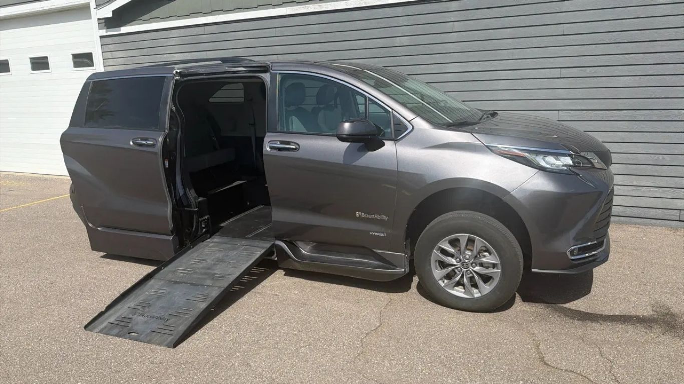 Gray minivan with side door open and wheelchair ramp deployed on pavement near a building