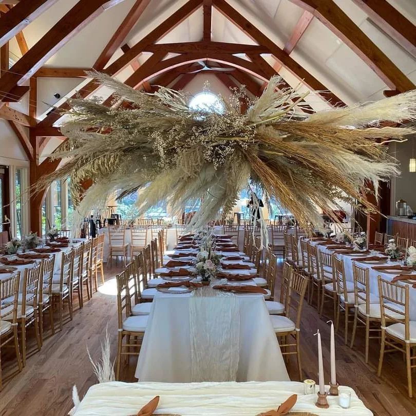 Wedding reception: long tables with white tablecloths, gold chairs, and an overhead floral arrangement in a wooden-beamed hall.